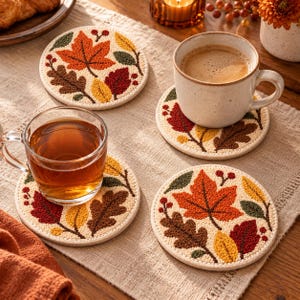 Puede incluir: Un juego de cuatro posavasos redondos con un patrón de hojas de otoño bordado y texturizado en tonos naranja, rojo, amarillo, marrón y verde oscuro sobre un fondo crema. Dos posavasos sostienen tazas de vidrio y cerámica con bebidas.