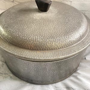 May include: A round, silver-colored metal pot with a matching lid, featuring a textured, hammered finish across the entire exterior surface. The lid is topped with a dark brown, faceted knob handle centered on the top surface.