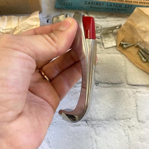 May include: A bright chromium cabinet latch set with a red plastic thumb piece. The latch is curved and has a sleek, vintage design. The packaging includes the text "Actual Size Bright Chromium Red Plastic Thumb Piece".