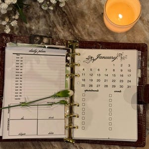 Può includere: Agenda in pelle marrone aperta con anelli dorati, con un piano giornaliero e un calendario di gennaio. Una candela accesa e fiori bianchi con steli verdi sono disposti attorno all'agenda su una superficie di marmo.