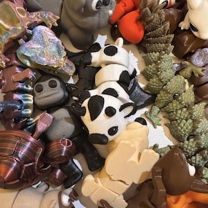 May include: A collection of colorful 3D-printed animal figurines, including a pug, cow, crab, and turtle. The figurines are in various colors, such as gray, white, black, red, and brown. The collection is diverse and features a variety of animal shapes.