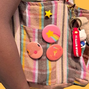 May include: A multi-coloured striped tote bag decorated with a yellow star pin, a pink pin featuring a rainbow star, a peach pin with a face, and a coral pin with banana text. A gold keychain with a red Hello Kitty strap is attached.