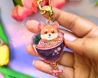 May include: An acrylic keychain featuring a cartoon Shiba Inu dog sitting inside a dark purple bowl of ramen with noodles, egg slices, and seaweed. The charm has a gold-colored cat-shaped clasp and a small dangling pink bow charm at the bottom.