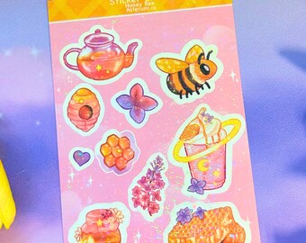 May include: A sticker sheet with a pink background featuring illustrations of a teapot, a honey bee, a beehive, a purple flower, a heart, a honeycomb cluster, a drink with a yellow ring, a floral sprig, and a piece of honeycomb with purple flowers.