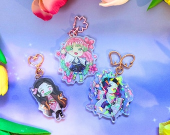 May include: Three acrylic keychains featuring anime-style characters. One shows a character with long pink and green hair, one with black hair and a bamboo muzzle, and one with dark hair and butterfly wings. Each has a gold-toned metal clasp.