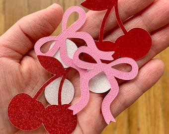 May include: A set of red and pink glitter paper cutouts of cherries with bows. The cherries are in the shape of a heart and have a white centre.