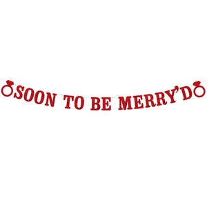 May include: A red glitter banner with the text "SOON TO BE MERRY'D" in capital letters. The banner is strung on a thin white string and features a ring design on each end. The banner is against a white background.