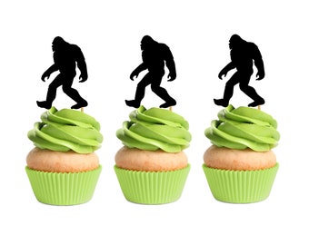 May include: Three green cupcakes with black silhouette Bigfoot cupcake toppers. The cupcakes have green frosting and are in white cupcake liners.