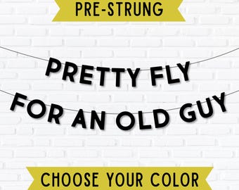 May include: A black banner with the text "PRETTY FLY FOR AN OLD GUY" strung across a white brick wall. The banner is pre-strung and includes the text "CHOOSE YOUR COLOR" in a yellow banner.