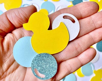 May include: A hand holding a variety of confetti for a baby shower. The confetti includes yellow rubber duckies, blue and white circles, and blue glitter circles.
