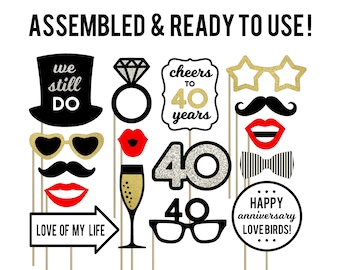May include: A set of 10 photo booth props for a 40th anniversary party. The props include a top hat with the words "we still do", a ring, a champagne glass, a pair of glasses, a bow tie, a moustache, a pair of lips, a star-shaped glasses, a sign that says "cheers to 40 years", and a sign that says "happy anniversary lovebirds!"