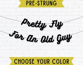 May include: Black banner with the phrase "Pretty Fly For An Old Guy" in a cursive font, strung between two lines. The banner is set against a white brick wall. The banner also includes the text "PRE-STRUNG" and "CHOOSE YOUR COLOR".