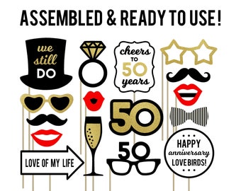 May include: A set of 10 gold and black photo booth props for a 50th anniversary party. The props include a top hat with the words "we still do", a pair of gold star-shaped sunglasses, a black bow tie, a red lip, a champagne glass, a gold ring, a gold number 50, a sign that says "love of my life", and a sign that says "happy anniversary love birds!"