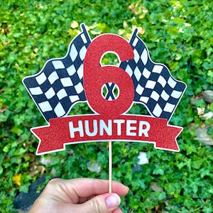 May include: Red glitter number six cake topper with black and white checkered racing flags and a red glitter banner that says "HUNTER".