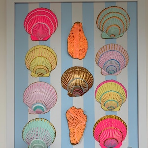 Puede incluir: Una pieza de arte mural enmarcada con un fondo de rayas verticales azul claro y blanco adornado con once conchas decorativas. Las conchas están pintadas en vibrante rosa neón, amarillo, verde menta, naranja y oro metálico con patrones de puntos.