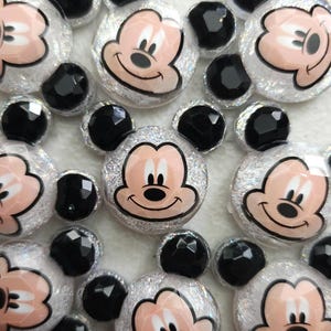 May include: A collection of flat-backed cabochons shaped like a cartoon mouse head. Each piece features a central face with a light peach skin tone and a silver glitter background, accented by two small, faceted, black circular ears on top.