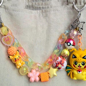 May include: A decorative bag charm featuring a pastel iridescent chain, assorted colorful beads, and miniature character figures including Pikachu and Charmander. The charm includes a yellow figure wearing a dragon costume and silver metal clasps.
