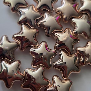 May include: A collection of small, five-pointed star-shaped beads with a smooth, reflective metallic rose gold finish. Each bead features a rounded, puffed design with a polished surface that catches light, creating bright highlights and reflections.