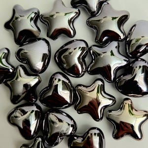 May include: A collection of shiny, silver-colored star and heart-shaped decorative beads. The beads have a reflective surface, creating a metallic appearance. The shapes are scattered across a white surface.