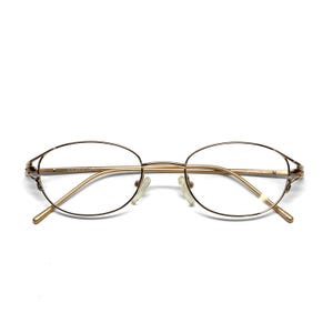 Puede incluir: Gafas ovaladas doradas con lentes transparentes. Las monturas tienen un diseño delicado y fino con sutiles detalles decorativos cerca de las patillas. Las gafas están sobre un fondo blanco.