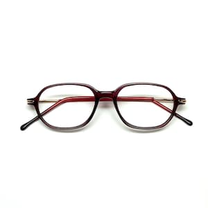 Op de afbeelding: Een bril met een bordeauxrood montuur en rode veren. De glazen zijn helder en het montuur heeft een afgeronde, achthoekige vorm. De veren hebben goudkleurige accenten en zwarte uiteinden. Op een witte achtergrond.