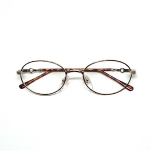 May include: Oval-framed spectacles with a thin, bronze-coloured metal frame. The temples have a decorative design, and the lenses are clear. The bridge and nose pads are a light bronze colour. The lenses are oval-shaped.