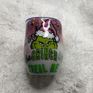 May include: A glitter-covered wine tumbler featuring a Grinch design. The Grinch is wearing a Santa hat and the text reads "I'm so the Grinch want to steal me" in green and pink. The tumbler has a pink and silver glitter design.