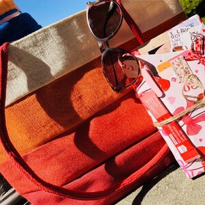 May include: A striped beach tote bag in orange, red, cream, and blue with a red rope handle. Sunglasses rest on top of the bag, next to a wrapped gift with a bow and a lip gloss.