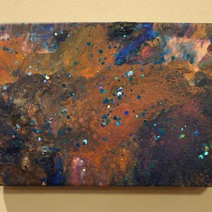 May include: Abstract acrylic painting on canvas. The artwork features a blend of warm and cool colors, including shades of brown, orange, and blue. Irregular shapes and splatters of glitter add texture and visual interest to the piece. The canvas is rectangular.