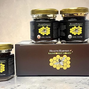 May include: Three glass jars of Health Harvest Rainforest Honey with gold lids, and a brown box with the same branding. The jars have black labels with a yellow honeycomb and bee graphic. The honey jars are 50g.