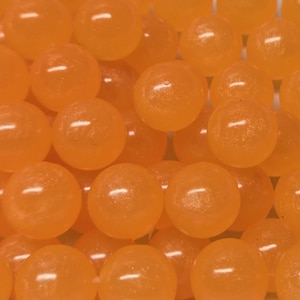 Könnte beinhalten: Eine Ansammlung zahlreicher kleiner, kugelförmiger, durchscheinender orangefarbener Perlen. Jede Perle hat eine glatte, polierte Oberfläche, die das Licht reflektiert und ein einheitliches Erscheinungsbild der in einem dichten Haufen angeordneten runden Objekte erzeugt.