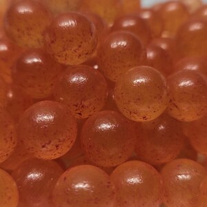Könnte beinhalten: Eine Ansammlung runder, durchscheinender orangefarbener Perlen mit gesprenkelter Textur. Jede Perle hat eine glatte, glänzende Oberfläche mit kleinen, dunkelorangefarbenen Einschlüssen, die gleichmäßig in der Kugelform verteilt sind.