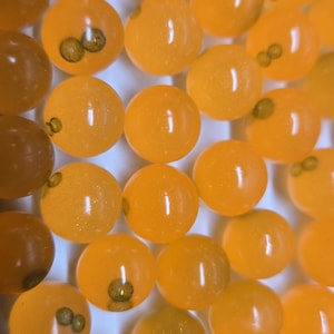 May include: A close-up shot of many translucent orange spheres, each with a small, dark inclusion. The spheres are closely packed, forming a textured, repeating pattern. The lighting accentuates the round shapes and the subtle colour variations.