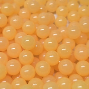 May include: A collection of numerous small, smooth, spherical beads in a uniform light peach or pale orange colour. Each bead features a glossy, reflective surface that catches the light, creating a dense, textured arrangement of round shapes.