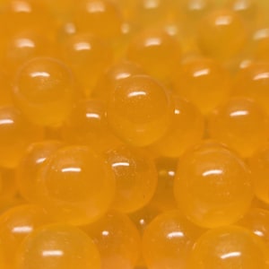 Könnte beinhalten: Eine Ansammlung kleiner, kugelförmiger, durchscheinender orangefarbener Perlen, die dicht beieinander liegen. Jede Perle hat eine glatte, glänzende Oberfläche, die das Licht reflektiert und ein einheitliches Erscheinungsbild erzeugt.