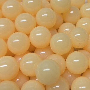 Könnte beinhalten: Eine Ansammlung zahlreicher glatter, runder, blass pfirsichfarbener Perlen. Jede Perle hat eine glänzende, reflektierende Oberfläche, die das Licht einfängt und kleine Glanzlichter auf den dicht beieinander liegenden, gleichmäßigen Kugeln erzeugt.
