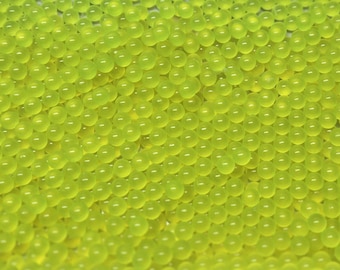 Può includere: Una vasta collezione di piccole perline sferiche e traslucide di colore giallo neon. Ogni perlina presenta una superficie liscia e lucida con una forma rotonda uniforme, creando uno strato denso e strutturato di sfere giallo brillante.