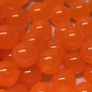 May include: A collection of small, round, translucent orange beads with a smooth, glossy finish. The beads are clustered together, creating a vibrant display of bright orange spheres that reflect light across their curved, polished surfaces.