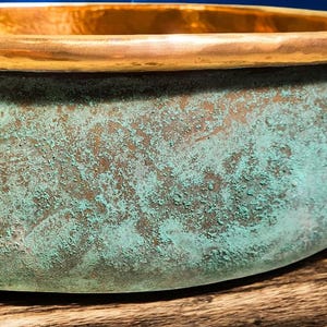 May include: Close-up of a copper bowl with a weathered, textured surface. The bowl's exterior features a turquoise patina over a bronze base, while the rim is a polished gold color. The bowl sits on a wooden surface.