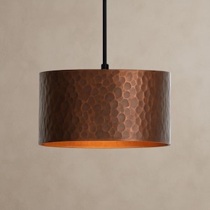 May include: A cylindrical pendant light fixture with a hammered copper shade. The shade has a textured surface and a warm, bronze color. The light hangs from a black cord.