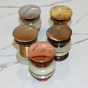 以下が含まれることがあります： 5つの装飾的な洗面台排水栓のコレクション。各栓は、グレー、ブラウン、銅色の丸い大理石模様のトップと、真鍮または銅色のベースが特徴です。栓は白とグレーの大理石の背景に配置されています。