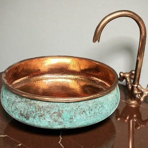 May include: A copper sink with a verdigris finish on the exterior and a polished interior. A matching copper faucet with cross handles is positioned to the right of the sink. The sink and faucet are on a brown surface.