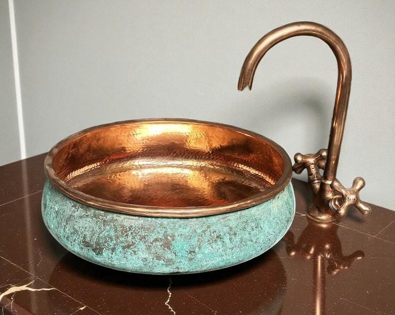 May include: A copper sink with a verdigris finish on the exterior and a polished interior. A matching copper faucet with cross handles is positioned to the right of the sink. The sink and faucet are on a brown surface.