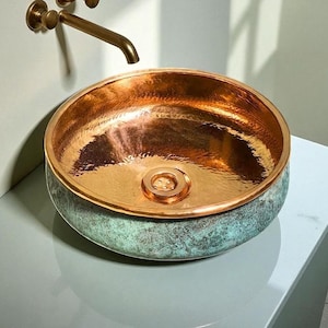 May include: A copper sink with a hammered interior and a verdigris exterior. The round basin has a polished copper rim and a central drain. A bronze faucet is mounted on the wall above the sink.