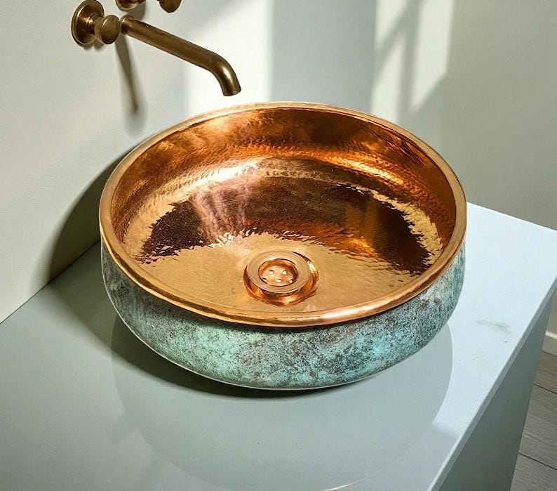 May include: A copper sink with a hammered interior and a verdigris exterior. The round basin has a polished copper rim and a central drain. A bronze faucet is mounted on the wall above the sink.