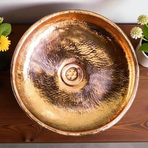 May include: A hammered copper sink with a round basin. The sink has a textured surface and a central drain. The copper has a warm, metallic sheen. The sink is displayed on a wooden surface.