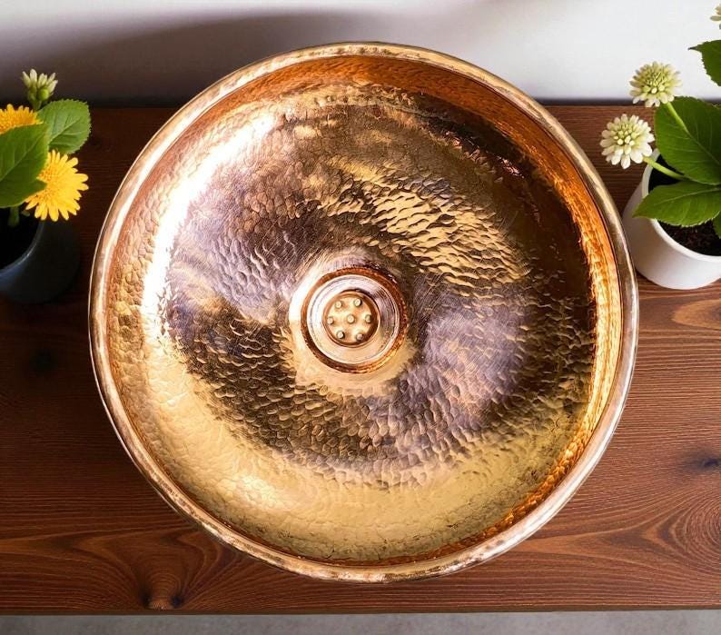 May include: A hammered copper sink with a round basin. The sink has a textured surface and a central drain. The copper has a warm, metallic sheen. The sink is displayed on a wooden surface.