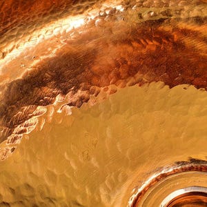 May include: Close-up of a hammered copper surface with a warm, golden-brown color. The texture shows a pattern of irregular, small indentations. The light reflects off the surface, creating highlights and shadows.