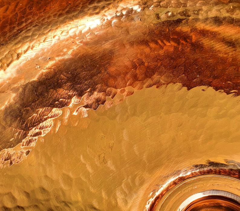 May include: Close-up of a hammered copper surface with a warm, golden-brown color. The texture shows a pattern of irregular, small indentations. The light reflects off the surface, creating highlights and shadows.