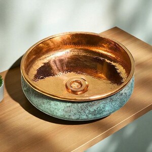 May include: A copper-colored, round vessel sink with a hammered interior and a verdigris exterior. The sink has a polished copper rim and a central drain. The sink is displayed on a wooden surface, showcasing its metallic sheen and unique color combination.
